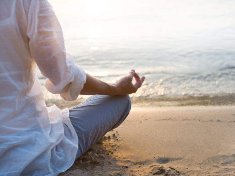 One (vertical) half of a person, from back, in white shirt sitting cross-legged at each of water on beach. Boost your attention with meditation.