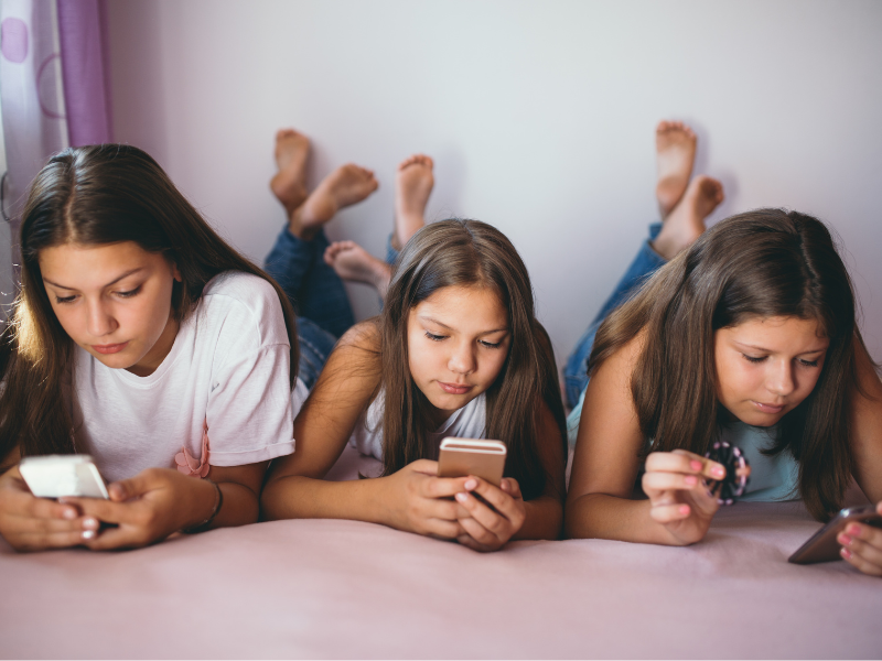 three young teen girls, lying prone next to each, looking at their mobile phones