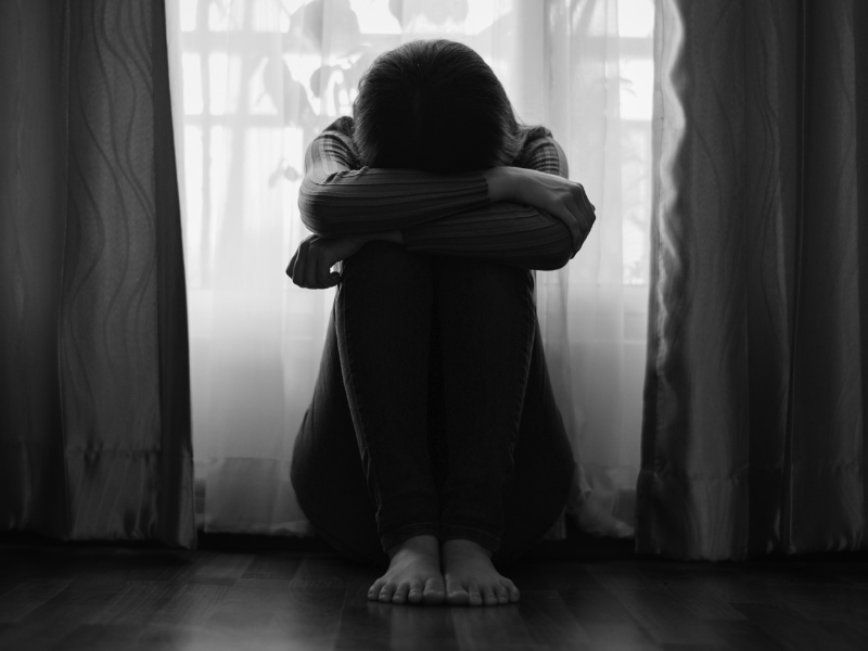 black and white photo of young person sitting on floor, knees bent, arms crossed over knees and head resting on arms. Background is curtains against a window.