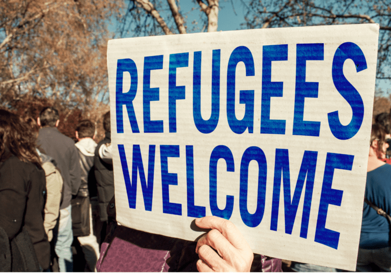 Amidst a crowd of people is a hand holding a Sign reading Refugees Welcome