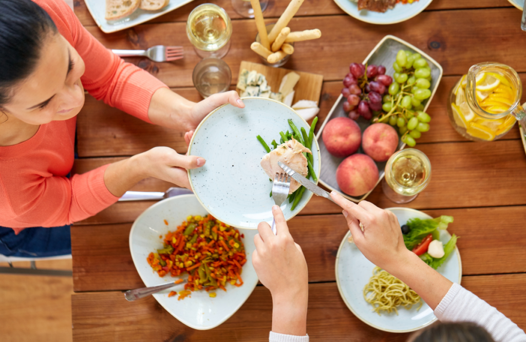 Bird's-eye view of 2 people sharing a meal. One person serving the other a plate with green beans and chicken. On the table is a fruit platter two glasses of white wine and two other plates of food. Plant-based proteins in diet.