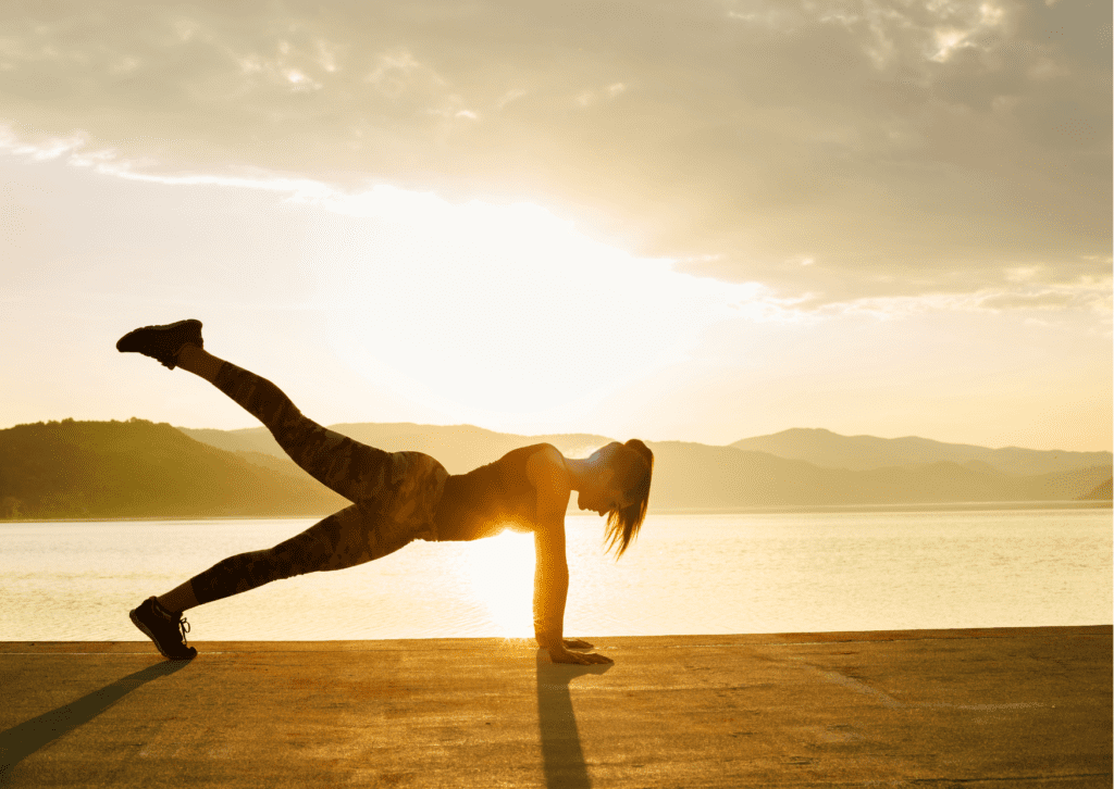 Woman doing pushup with one leg in the air. She is silhouetted against a sunset, with a lake and hills in the background.