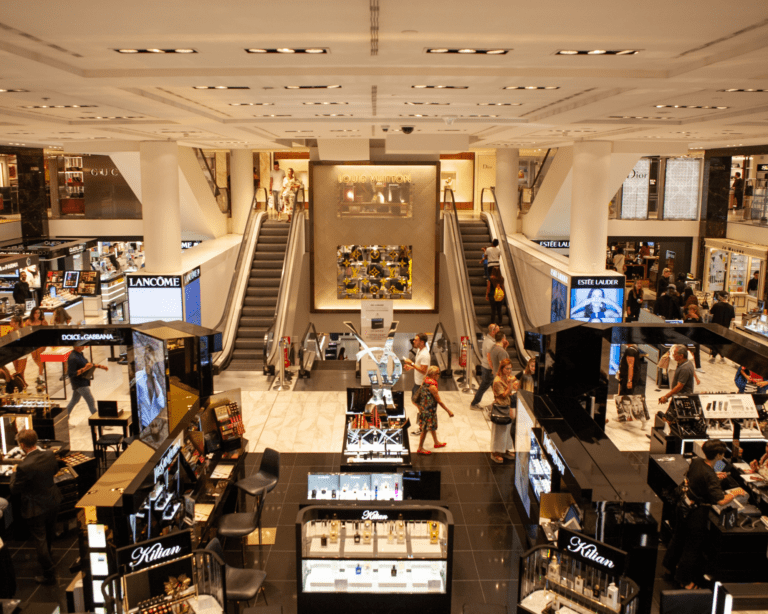 Photo of people walking around a department store showing multiple brand names, products and two escalators divided by a large ad for Louis Vuitton.