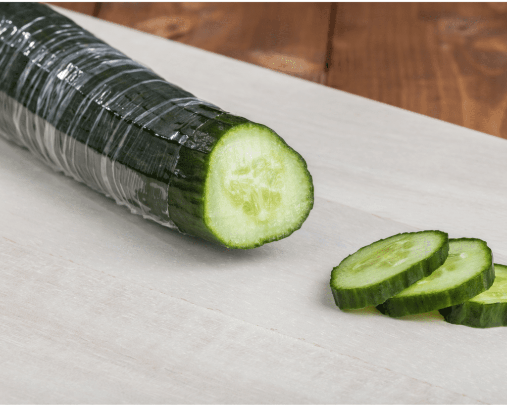 A plastic wrapped cucumber on bench with three slices cut from end.