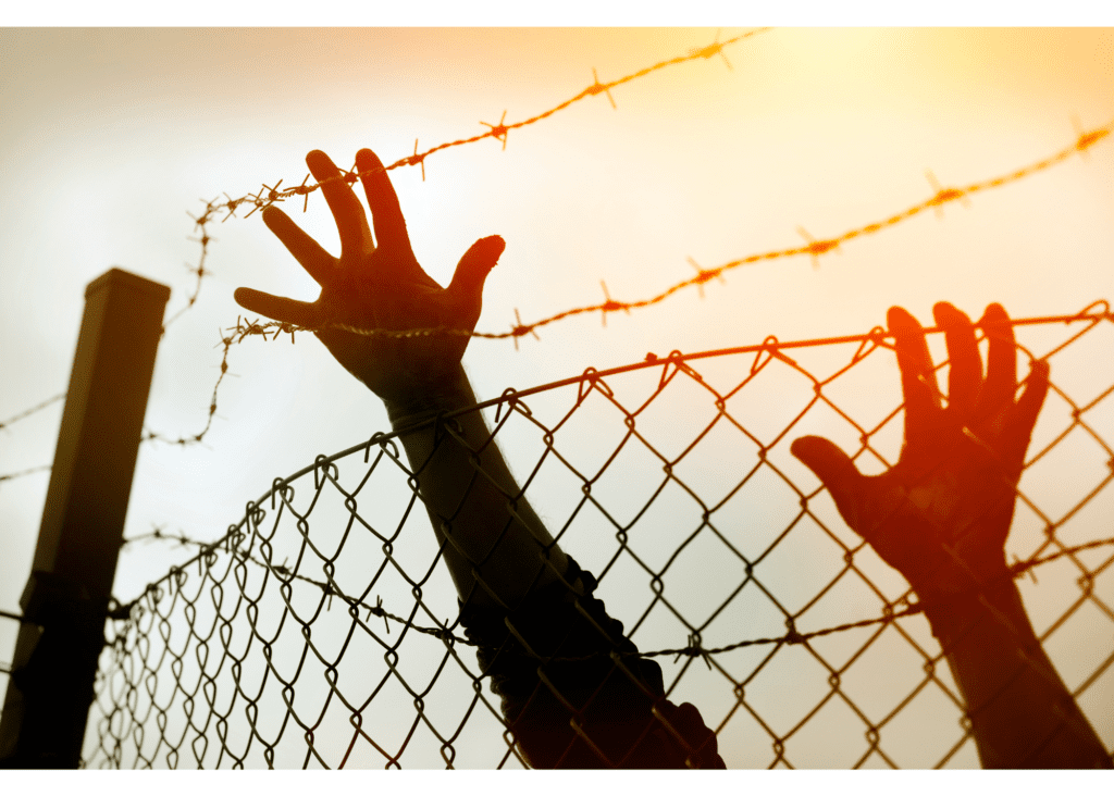 Two hands, silhouetted by the sun, tying to reach over a barbed wire fence.