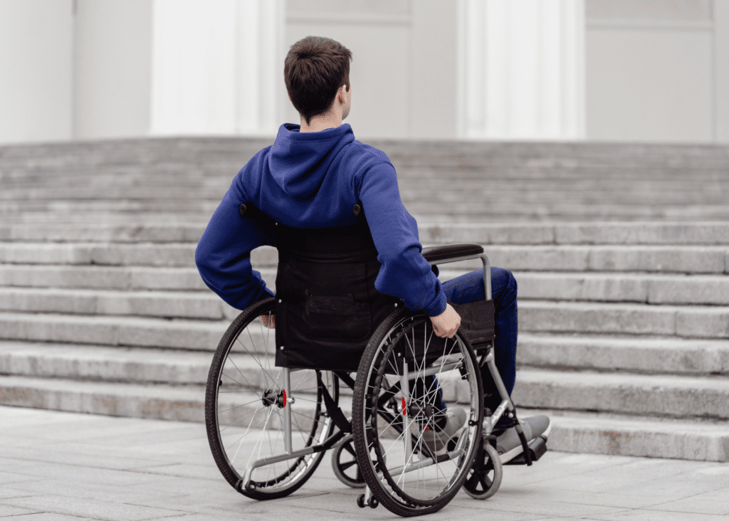 Back of young man wearing a blue hoodie and sitting in a wheelchair at the bottom of a flight of concrete steps.