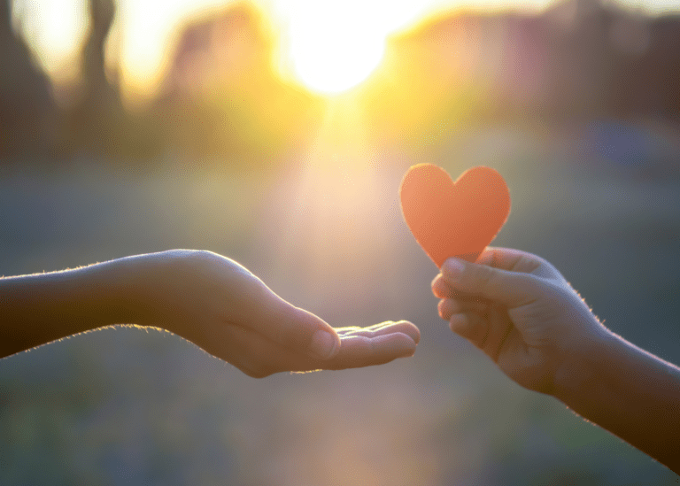 Photo of a hand holding up a red love heart to another hand receiving with sunset in background.