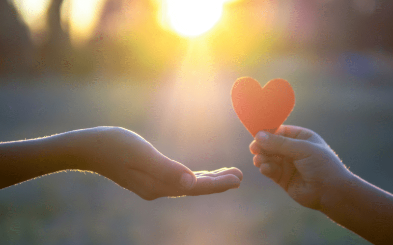 Photo of a hand holding up a red love heart to another hand receiving with sunset in background.