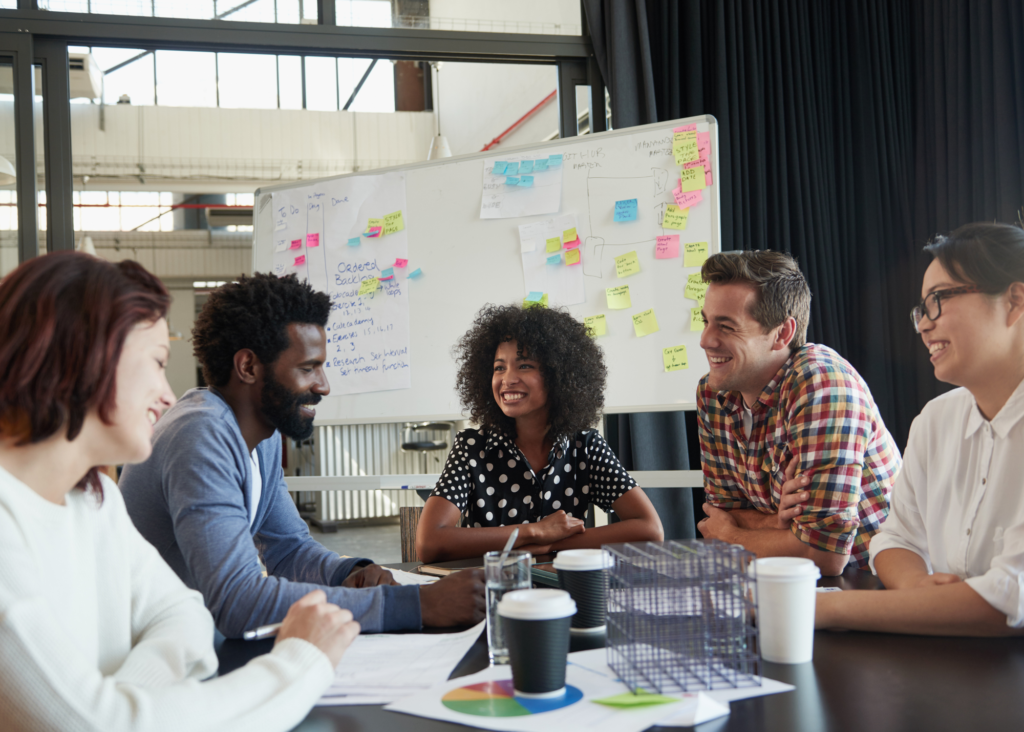 A group of five men and women sitting around a desk with papers and coffee cups on. A whiteboard with coloured sticky notes and writing is behind them.