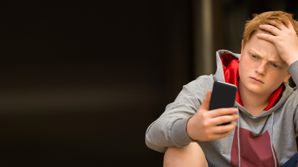 A young boy sitting looking perplexedly at his phone. Displaying that we need to redefine masculinity in the age of the internet.