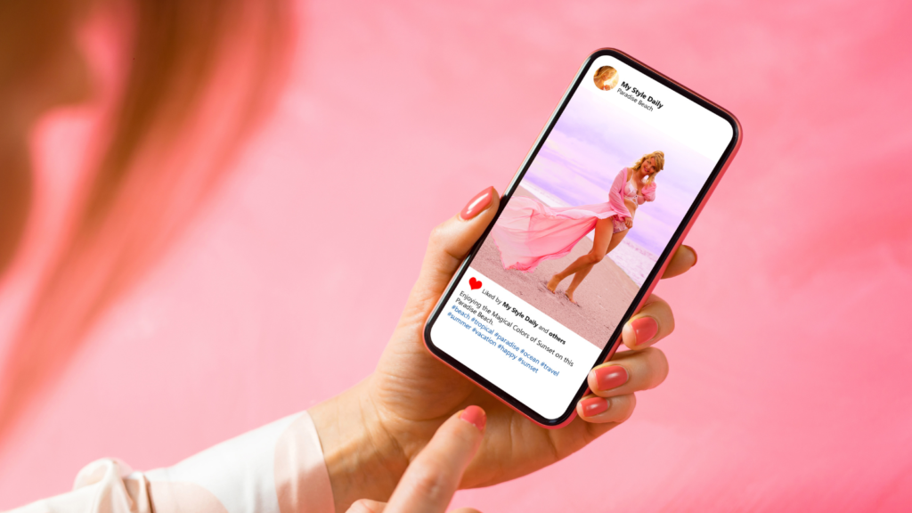 A female hand holding a mobile phone displaying social media post of a woman on a beach, against a pink background.