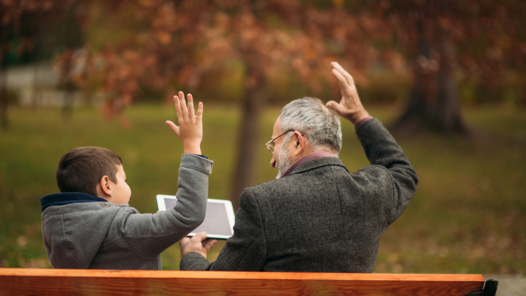 The rear view of an older man and a young boy sitting on a park bench together each with a hand in the air.