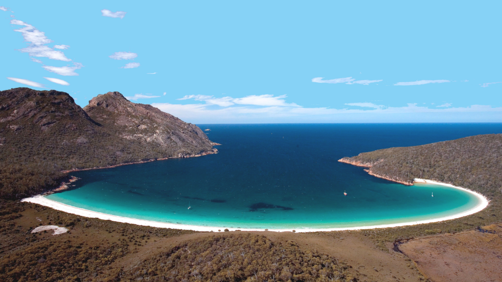 A view of Wine-Glass Bay Tasmania—a blue ocean bay with white sand and surrounded by hills of bushland.