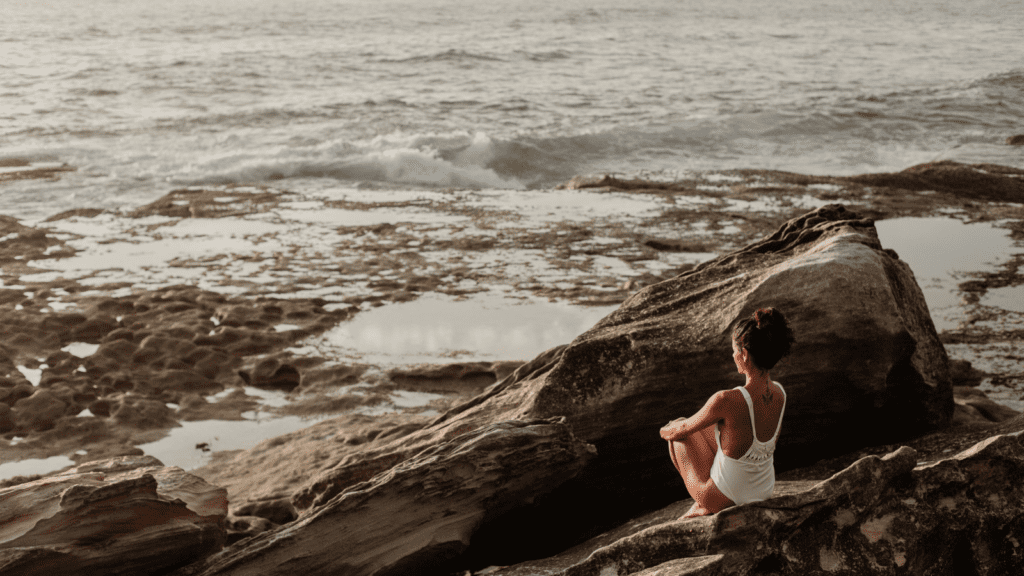 Woman on rocks looking out at ocean, prioritising self-care and sustaining physical, mental, and emotional well-being.